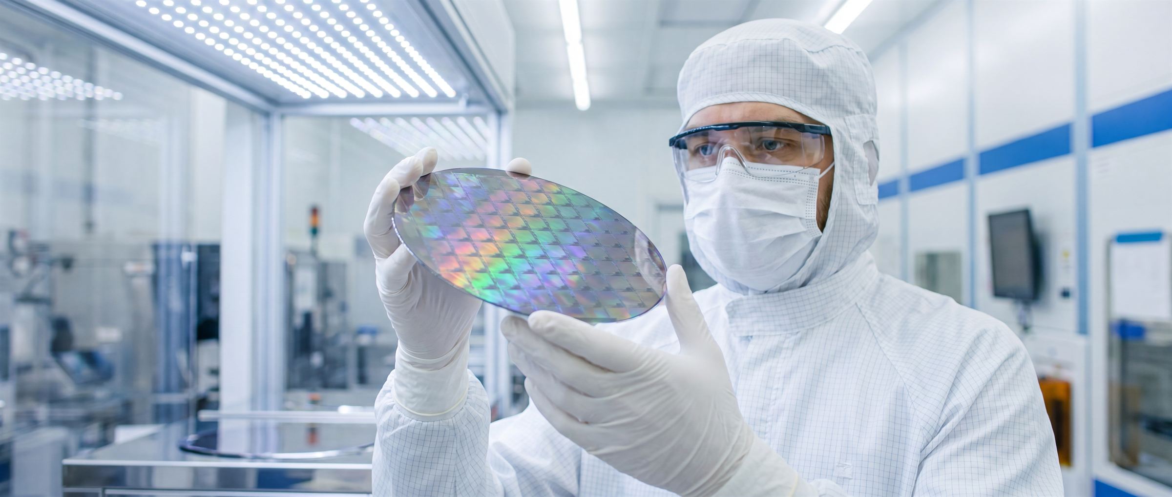 An engineer in cleanroom garments inspecting a patterned silicon wafer.